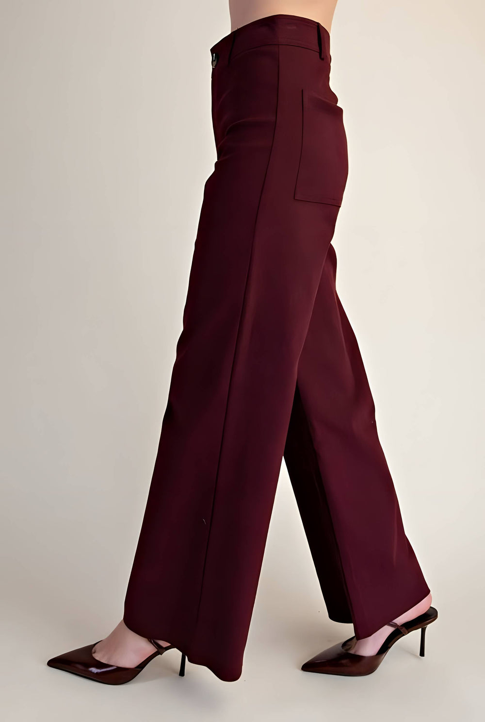 A person wearing high-waisted, wide-leg trousers in a deep burgundy or maroon shade, paired with dark brown pointed-toe heels, shown from the waist down against a plain light beige studio background.