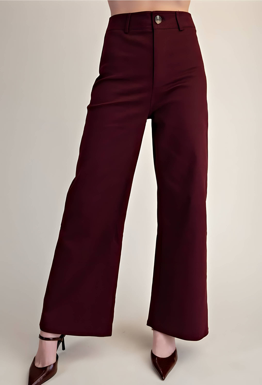 A model is wearing high-waisted, wide-leg trousers in a deep burgundy color, paired with dark brown pointed-toe heels, against a plain light beige studio background.
