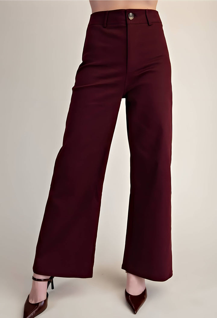 A model is wearing high-waisted, wide-leg trousers in a deep burgundy color, paired with dark brown pointed-toe heels, against a plain light beige studio background.