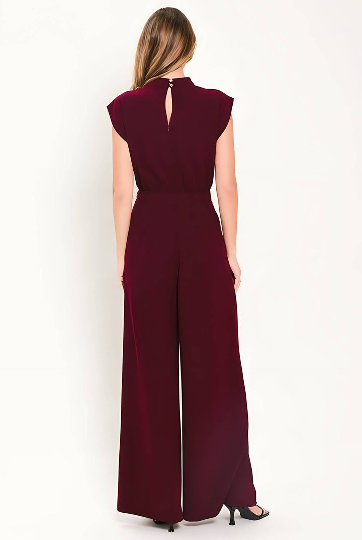 A woman viewed from the back wearing a sleeveless burgundy jumpsuit with a high neckline, keyhole back detail, and wide legs, standing against a plain white studio background.