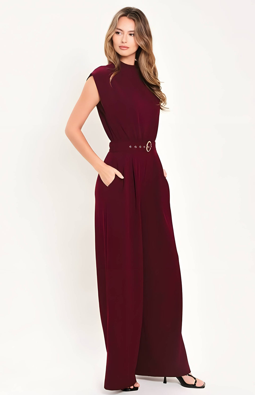 A woman modeling a sleeveless burgundy jumpsuit with a matching belt featuring a gold-tone buckle, set against a plain white studio background.