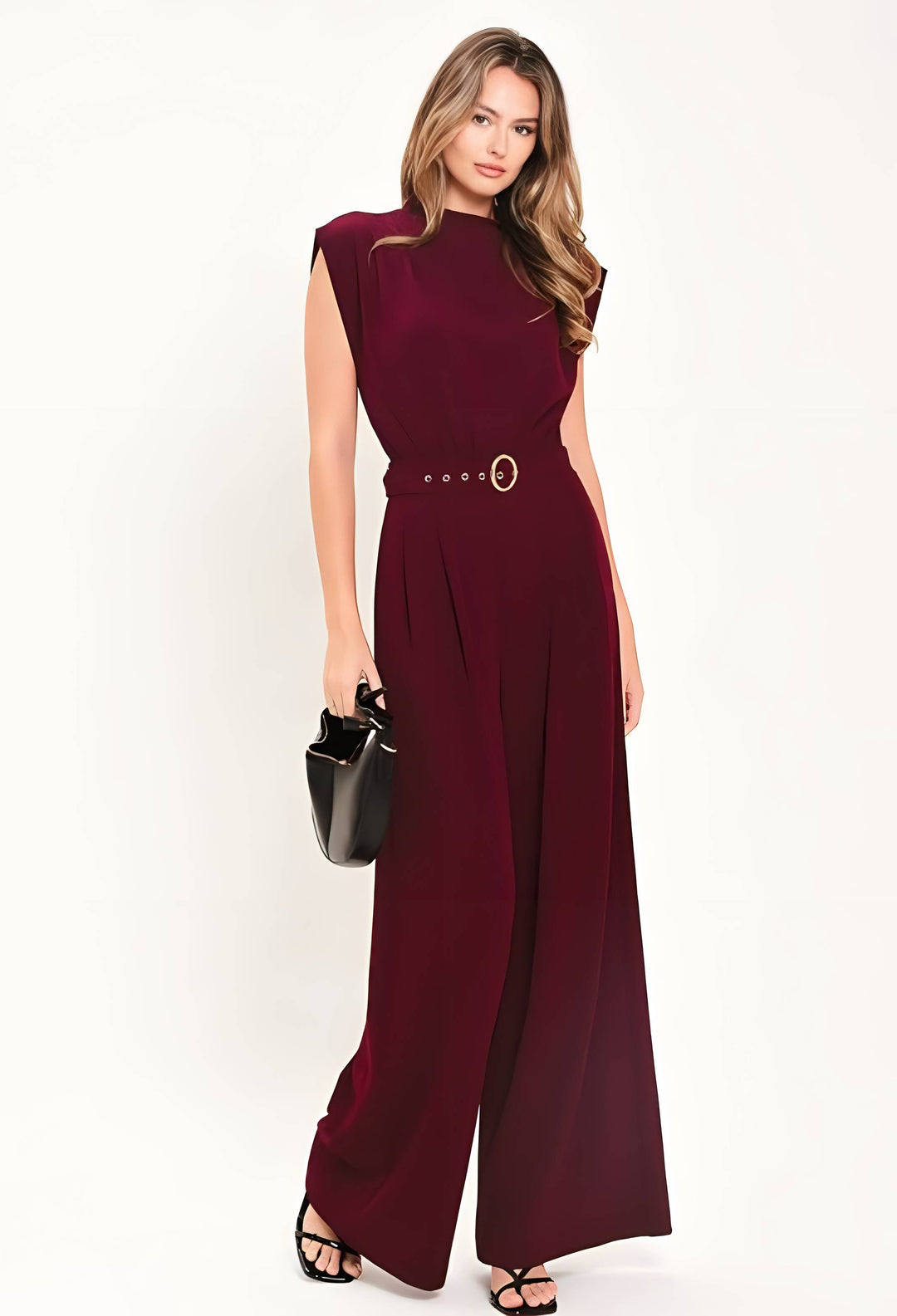 A woman modeling a sleeveless deep burgundy wide-leg jumpsuit with a matching belt and gold-tone buckle, holding a small black handbag against a plain white studio background.