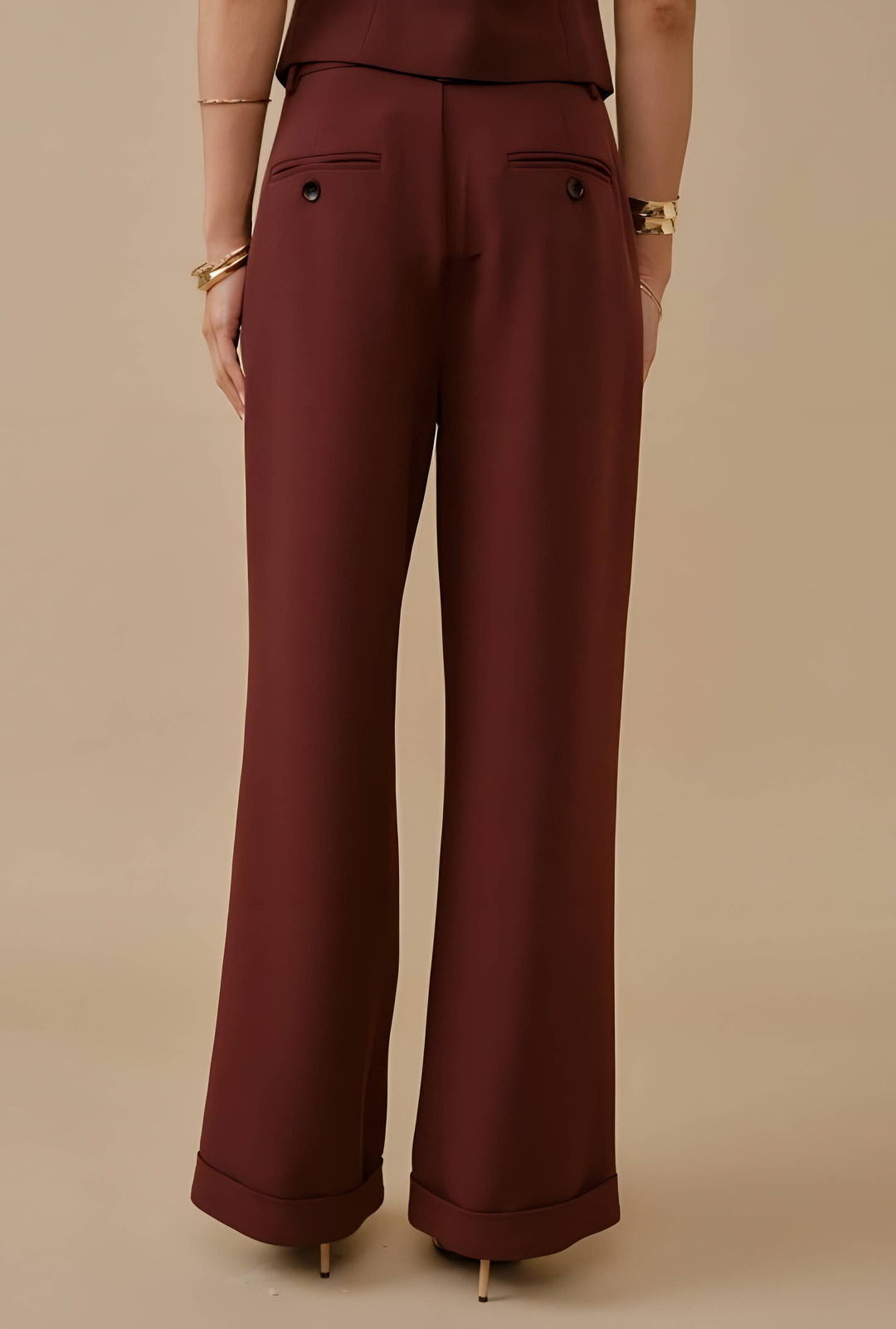 A person shown from the back wearing burgundy wide-leg trousers with a small button detail on the back pockets, paired with a matching top against a solid beige studio background.