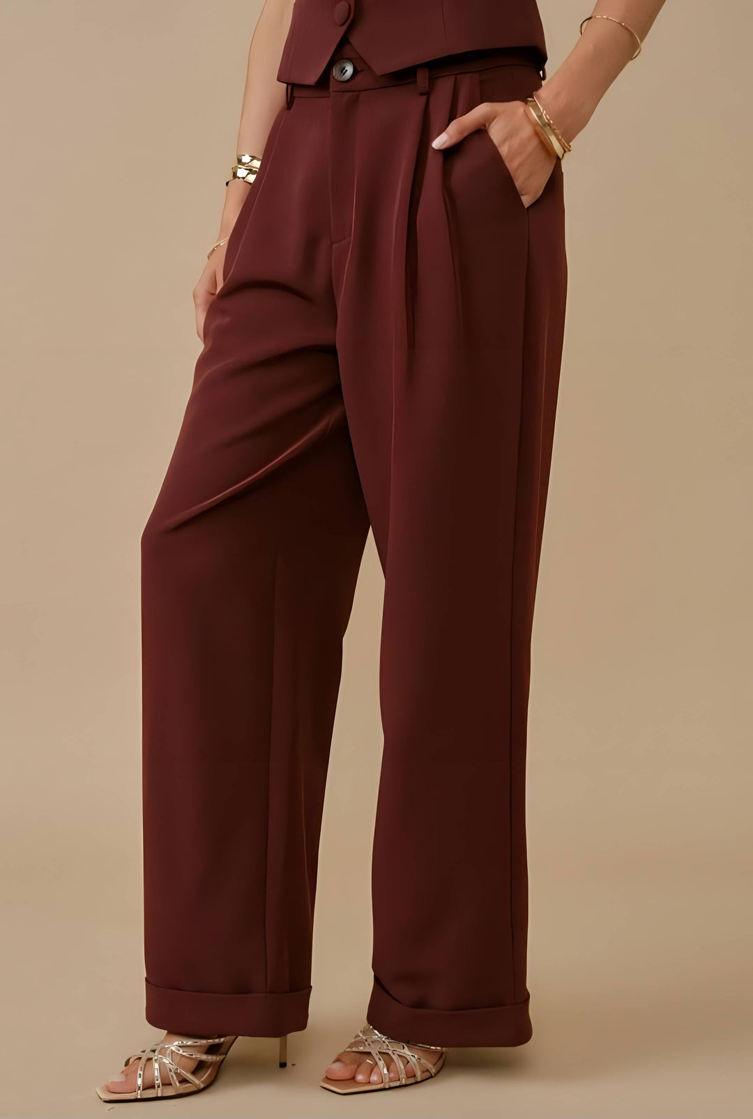 A person wearing high-waisted, wide-leg trousers in a deep burgundy or maroon shade, paired with metallic heeled sandals and gold bracelets, against a solid beige studio background.