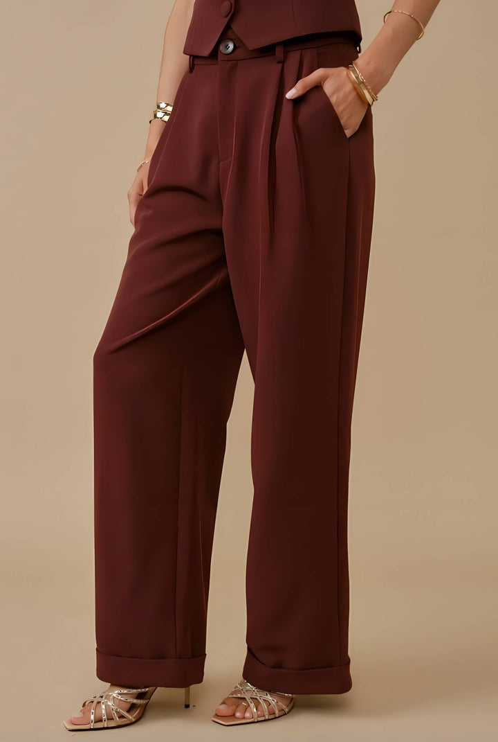 A person wearing high-waisted, wide-leg trousers in a deep burgundy or maroon shade, paired with metallic heeled sandals and gold bracelets, against a solid beige studio background.