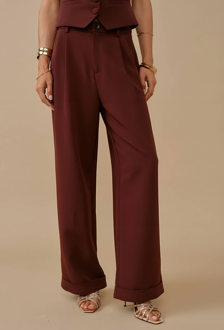 A model wearing a pair of high-waisted, wide-leg trousers in a deep burgundy or maroon shade, paired with a matching cropped vest and metallic sandals, set against a solid beige studio background.