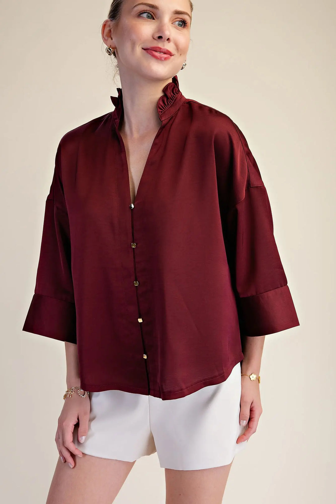 Model wearing a burgundy button down top with 3/4 sleeves and a ruffle neck with white shorts