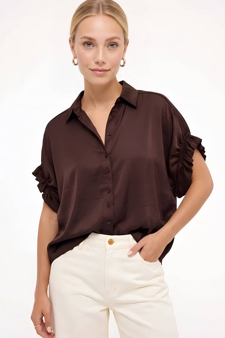 A woman modeling a dark brown satin-like button-up blouse with ruffled short sleeves, paired with cream-colored pants, against a plain white studio background.