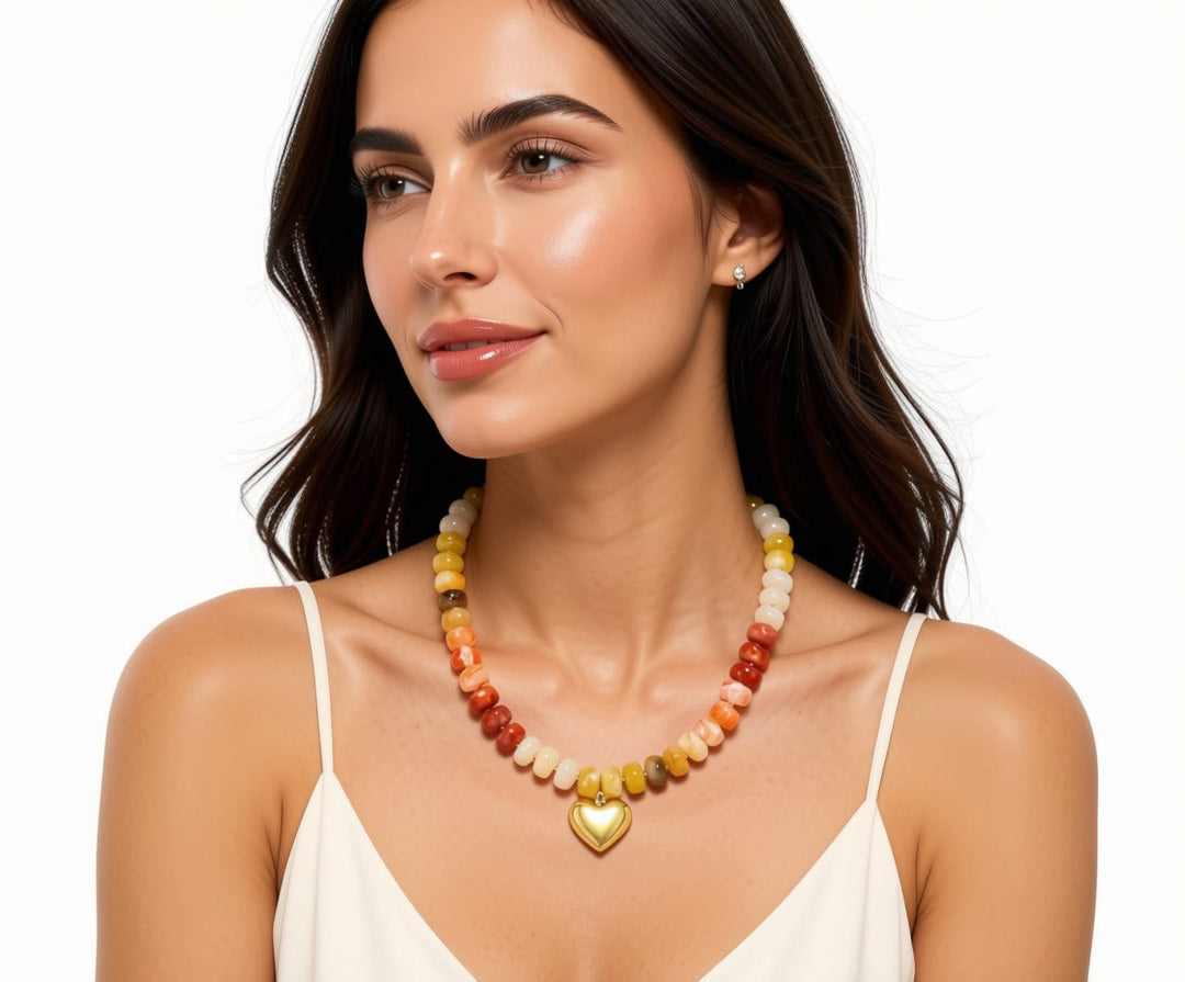 A woman wearing a colorful beaded necklace with a gold heart-shaped pendant, paired with a white spaghetti-strap top against a plain white background.