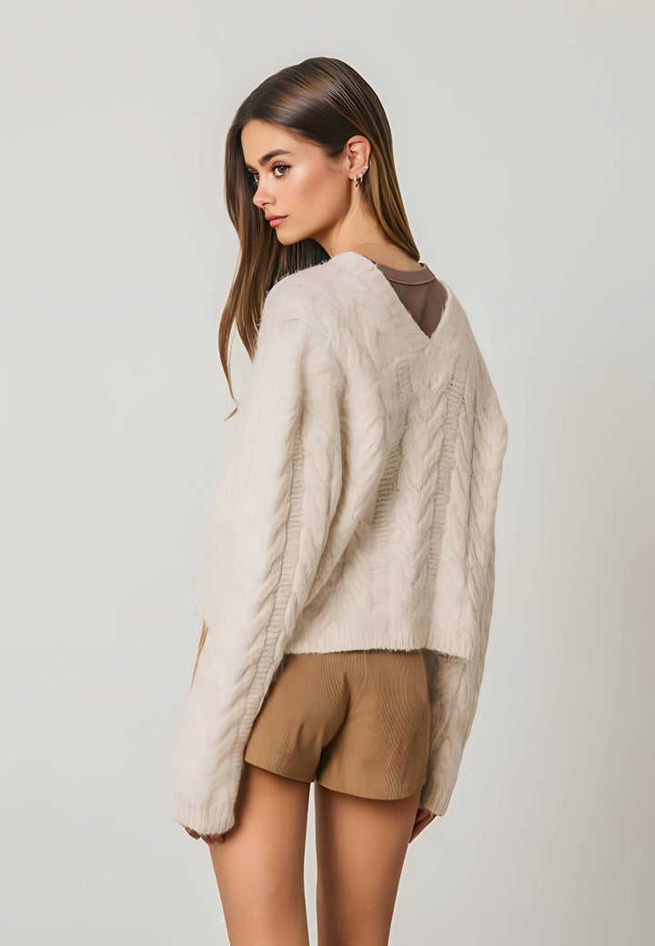A woman shown from the back wearing a cream cable-knit cropped cardigan over a brown top, paired with tan shorts, against a light gray studio background.