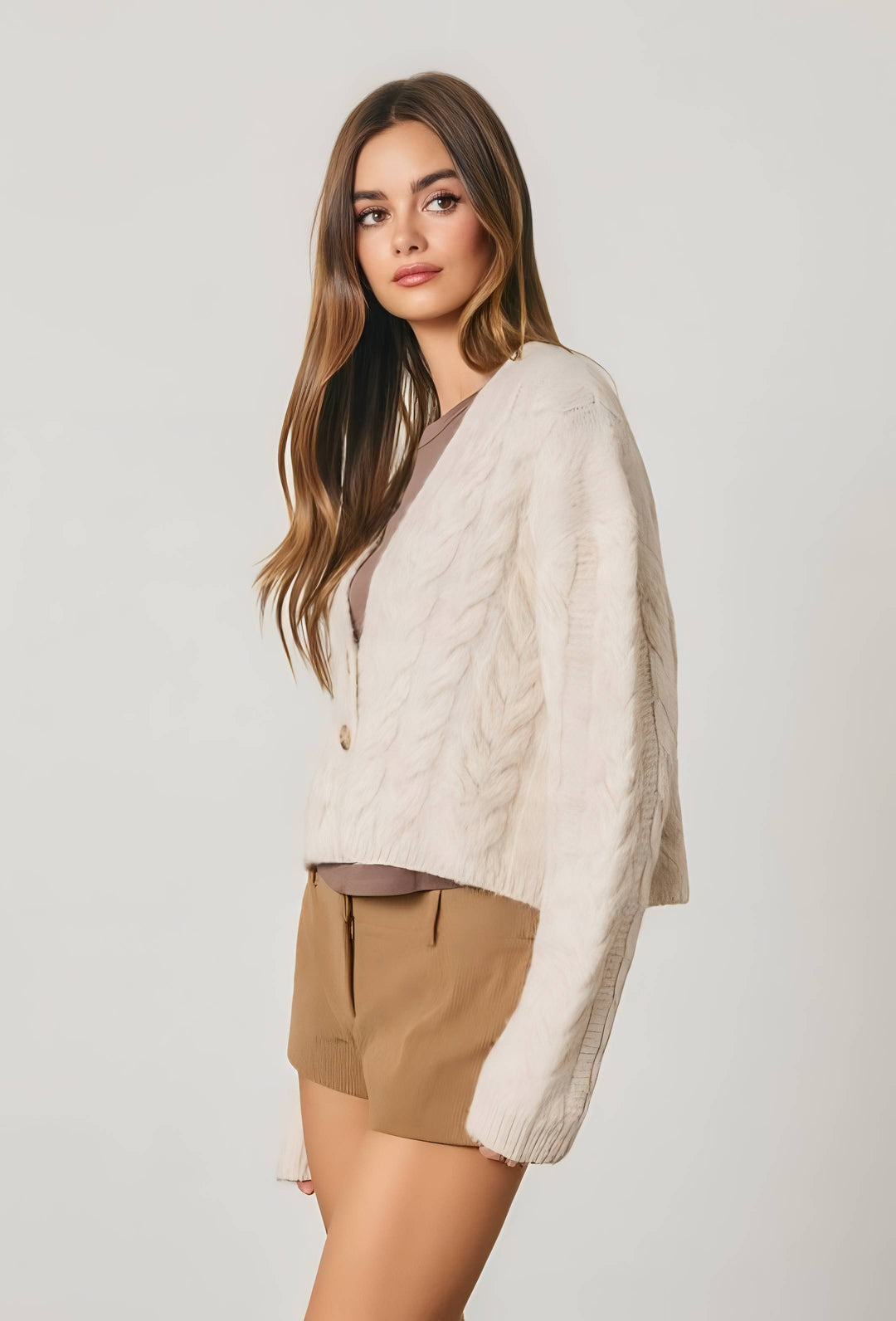 A woman modeling a cream cable-knit cardigan sweater over a taupe top and tan shorts, photographed against a plain light gray studio background.