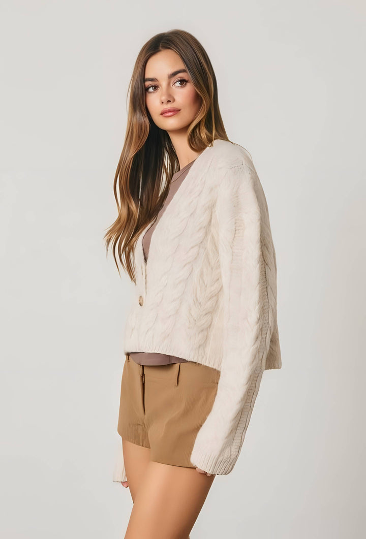 A woman modeling a cream cable-knit cardigan sweater over a taupe top and tan shorts, photographed against a plain light gray studio background.