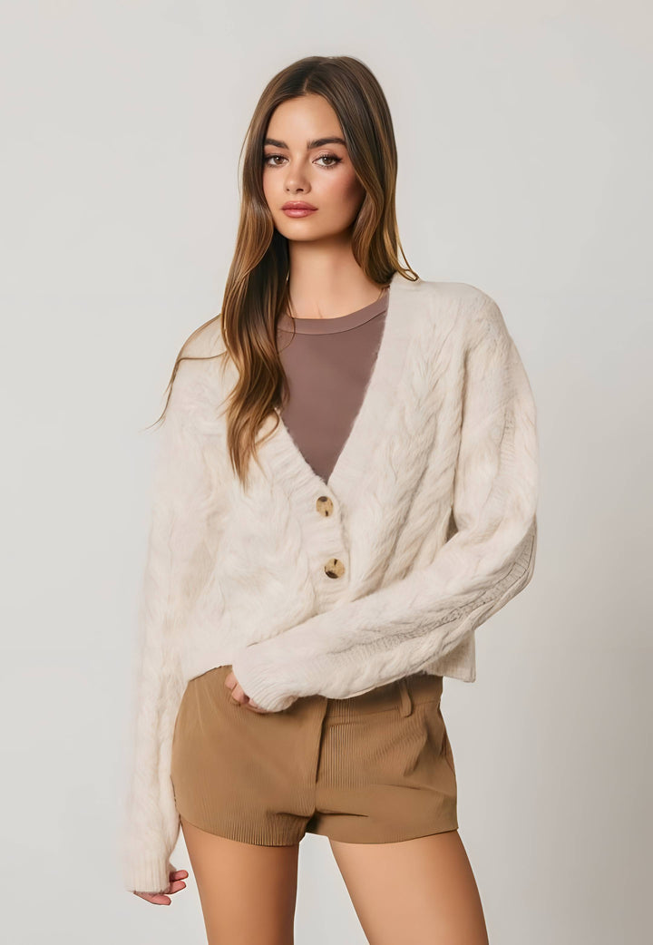 A woman modeling a cream cable-knit cardigan with gold buttons over a mauve-brown top and tan shorts, set against a plain light gray studio background.