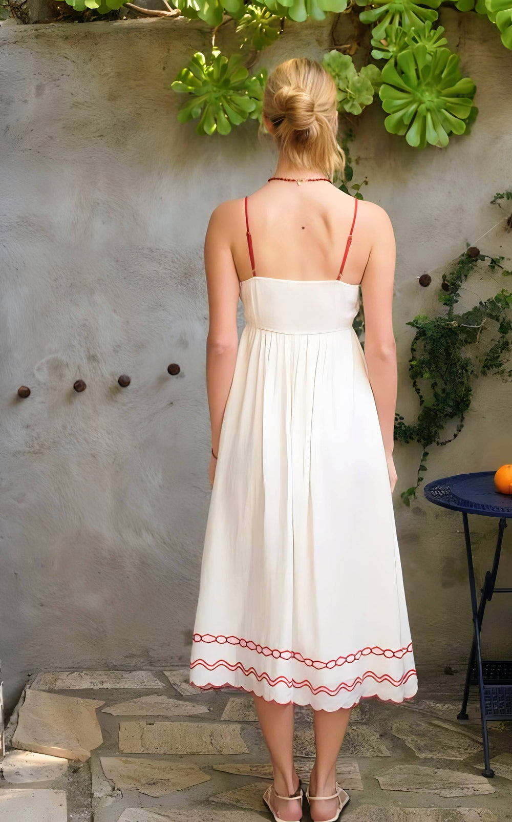 Woman in a white dress with red embroidery standing against a textured wall with greenery.