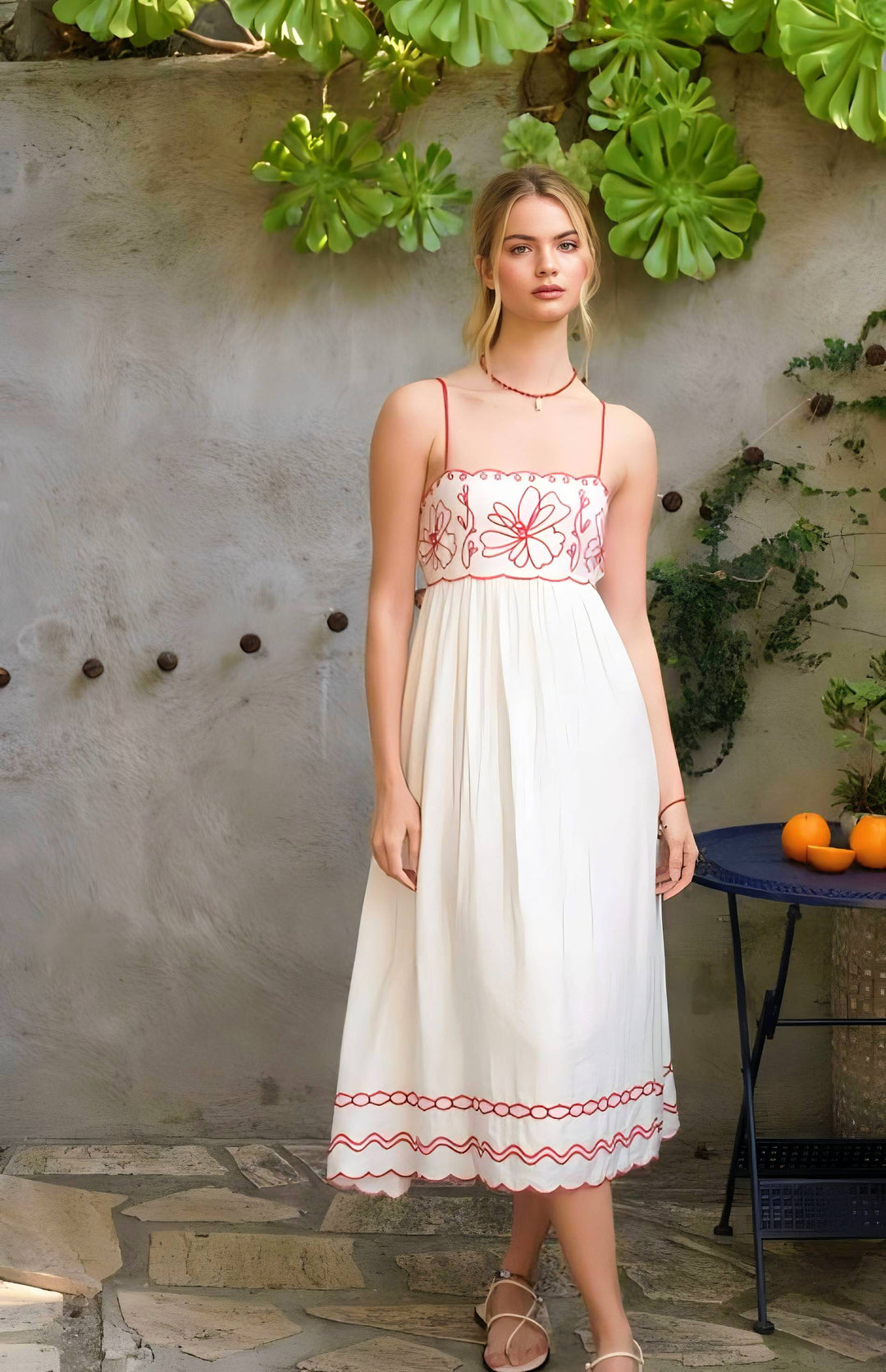 Woman wearing a white dress with red embroidery standing in front of a stone wall with green plants.