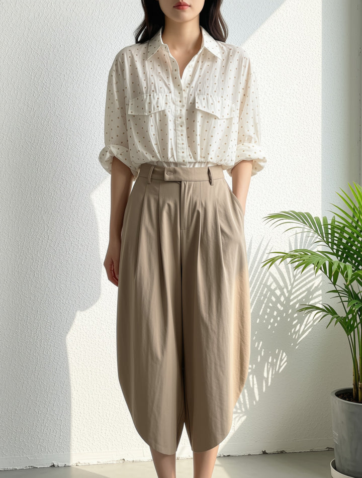 A woman wearing a white short-sleeve button-up shirt with a subtle black dot pattern and beige high-waisted wide-leg pants, standing against a white wall with a potted plant to the side.