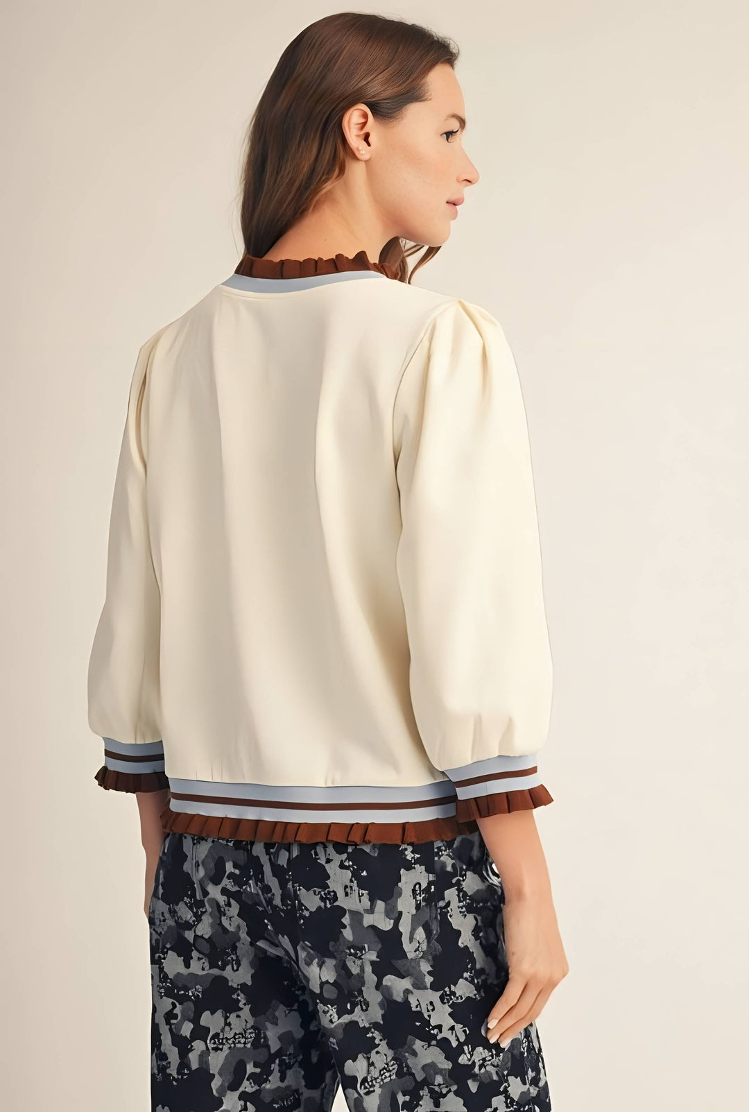 A woman shown from the back wearing a cream-colored blouse with brown ruffle trim at the collar, cuffs, and hem, paired with dark patterned trousers, against a plain beige studio background.
