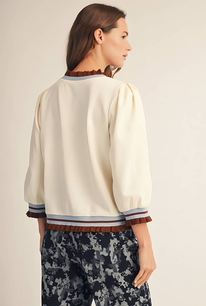 A woman shown from the back wearing a cream-colored blouse with brown ruffle trim at the collar, cuffs, and hem, paired with dark patterned trousers, against a plain beige studio background.