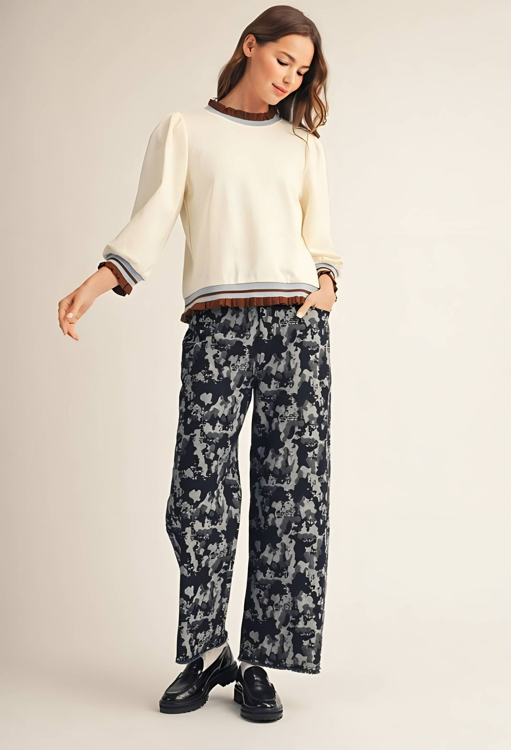 A woman modeling a cream-colored crewneck sweater with striped cuffs and hem, paired with dark patterned wide-leg trousers and black loafers, against a solid light beige background.