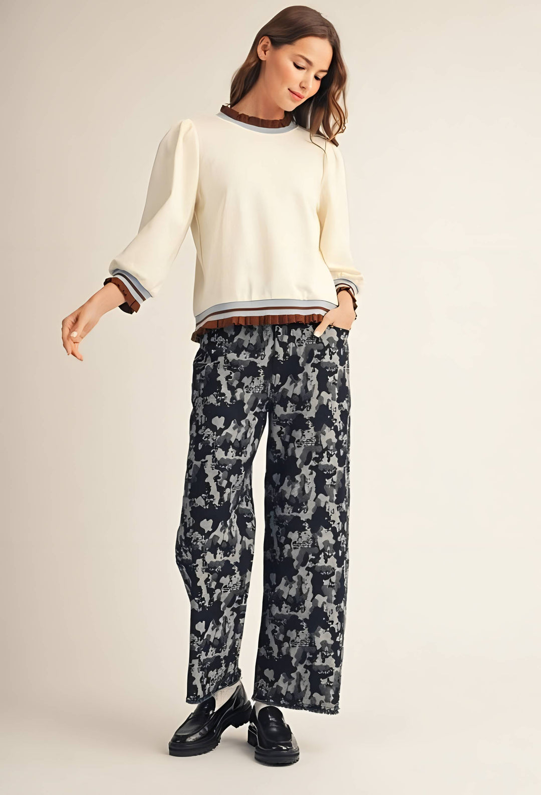 A woman modeling a cream-colored crewneck sweater with striped cuffs and hem, paired with dark patterned wide-leg trousers and black loafers, against a solid light beige background.