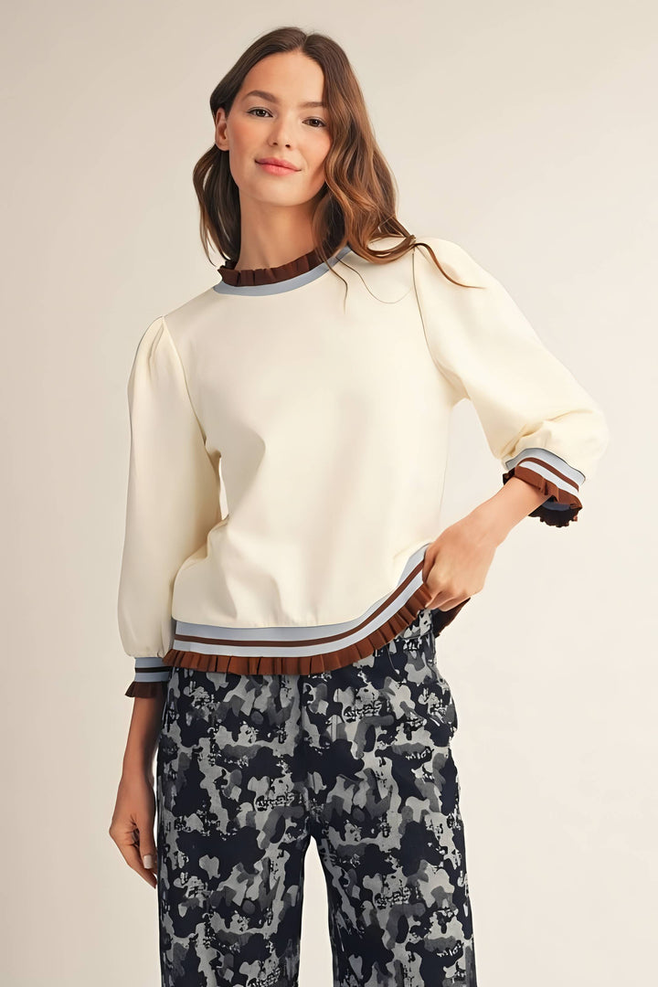 A woman modeling a cream-colored knit sweater with brown and gray striped ribbed trim at the crew neck, cuffs, and hem, paired with dark patterned pants, against a solid light beige background.
