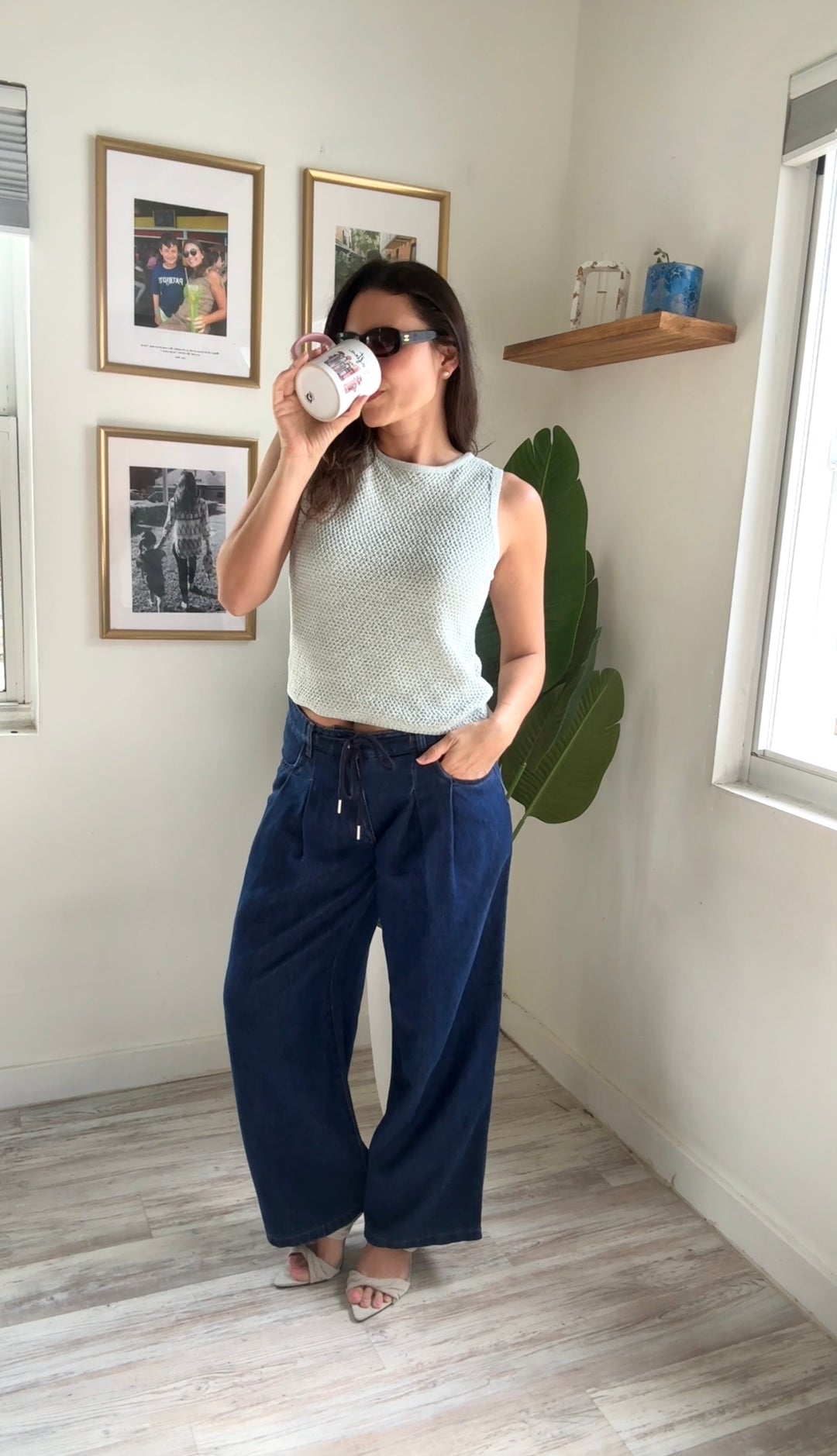Woman in a room with framed pictures and a plant, wearing a sleeveless top and wide-leg pants.