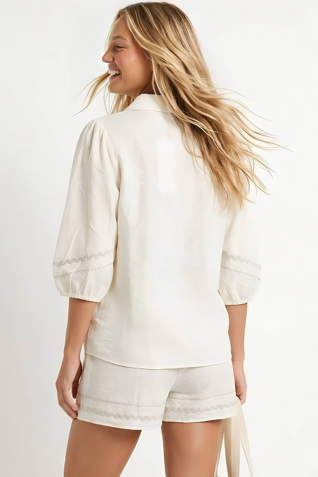 A woman shown from the back wearing a cream-colored puff-sleeve blouse and matching cream shorts with delicate lace trim, set against a plain white studio background.