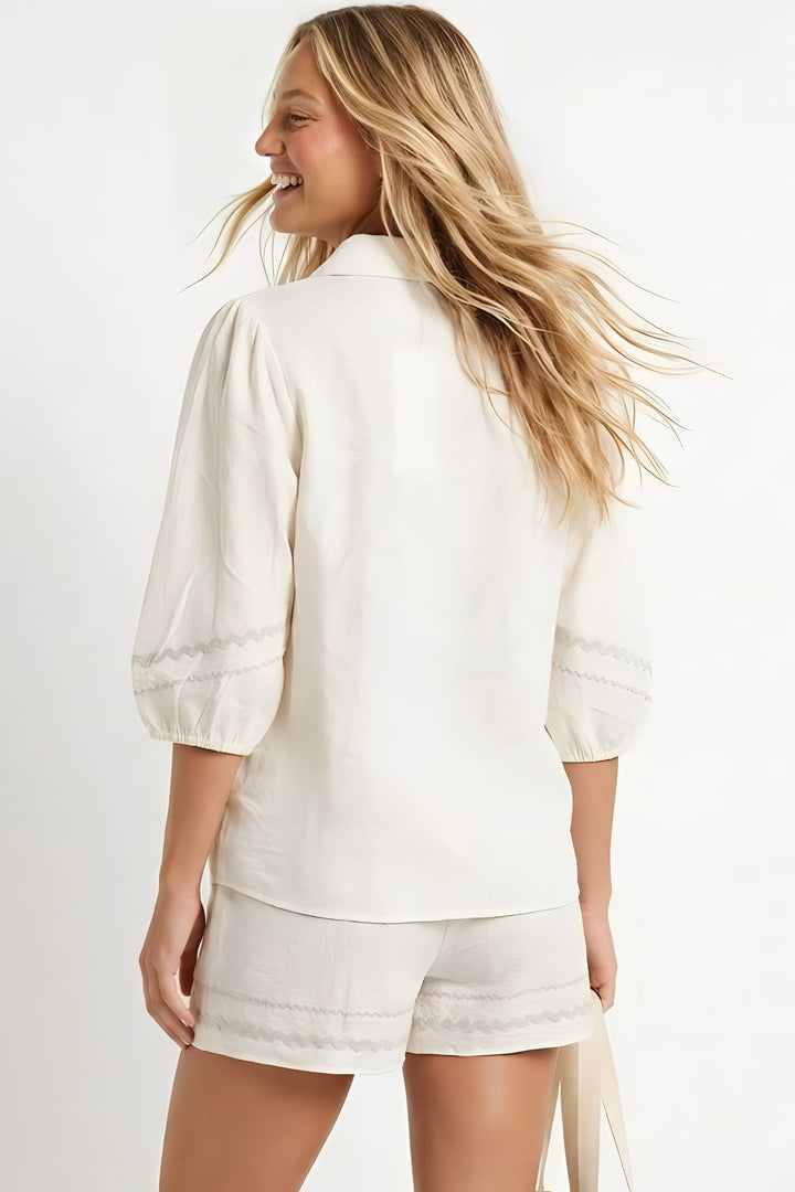 A woman shown from the back wearing a cream-colored puff-sleeve blouse and matching cream shorts with delicate lace trim, set against a plain white studio background.
