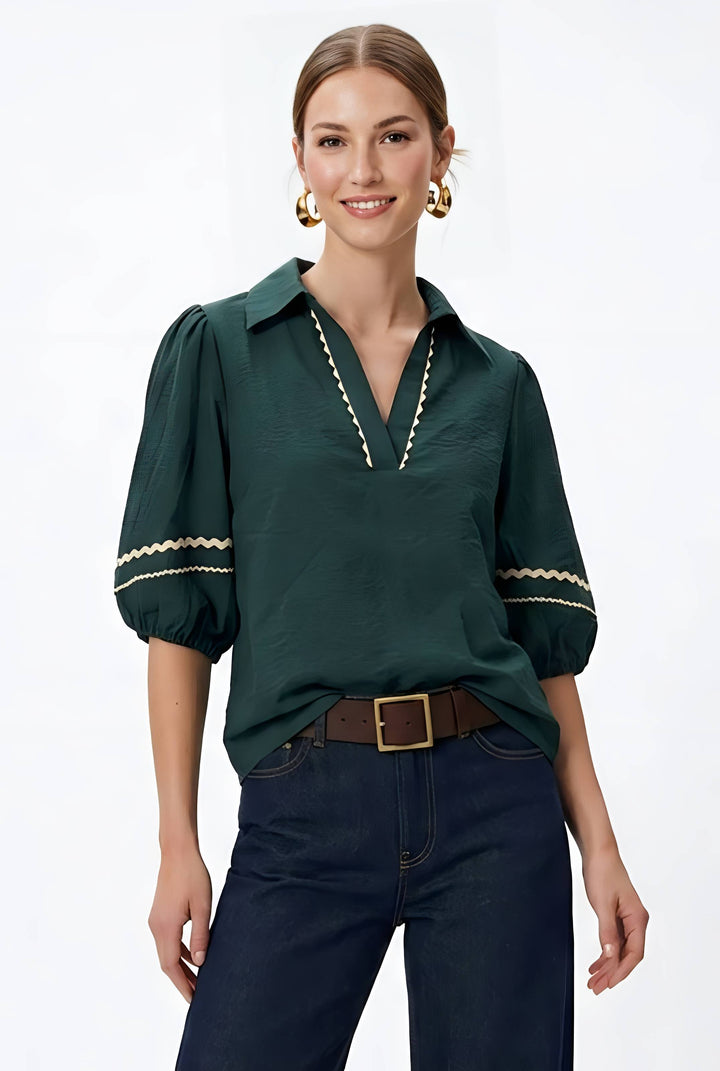 A woman modeling a dark green short-sleeve blouse with gold embroidered trim along the collar, placket, and sleeve cuffs, paired with dark blue jeans and a brown belt against a plain white studio background.