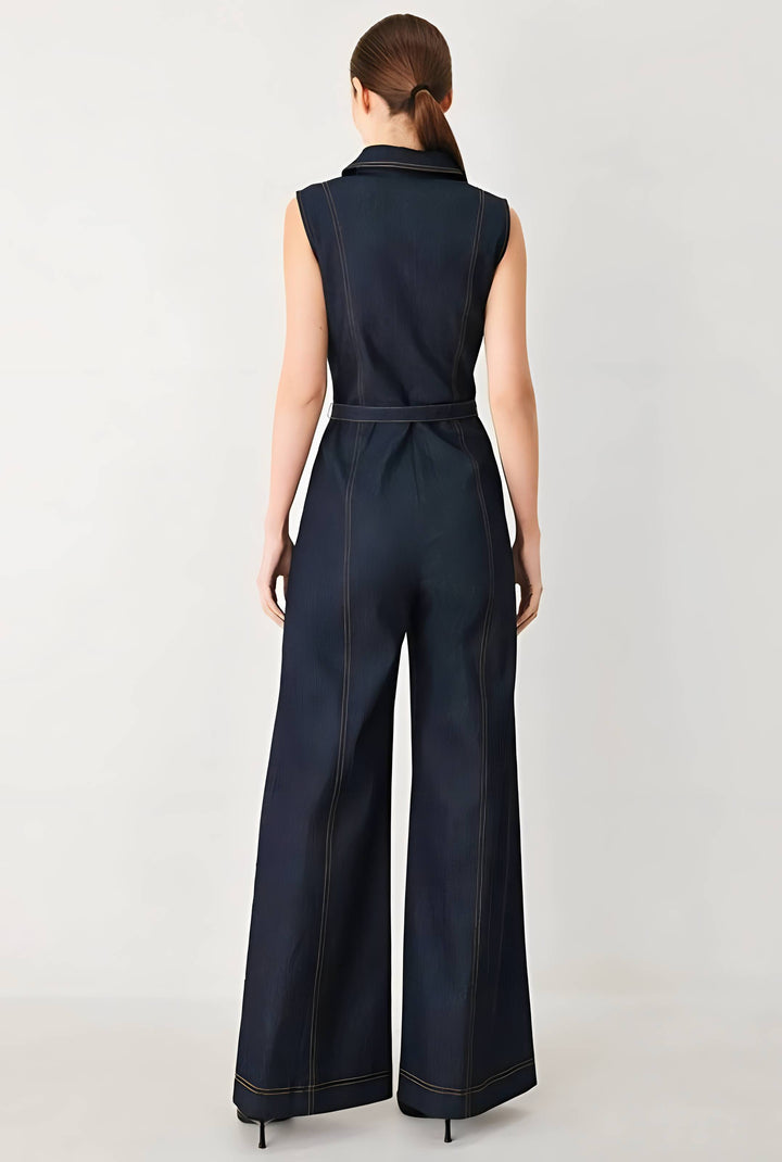 A woman viewed from the back wearing a sleeveless dark navy wide-leg jumpsuit with a matching waist tie, photographed against a light gray studio background.