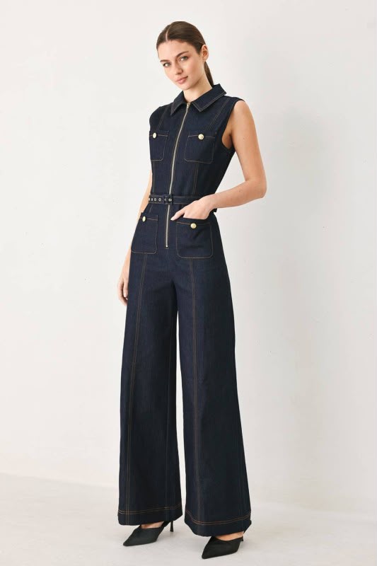 A woman viewed from the back wearing a sleeveless dark navy wide-leg jumpsuit with a matching waist tie, photographed against a light gray studio background.
