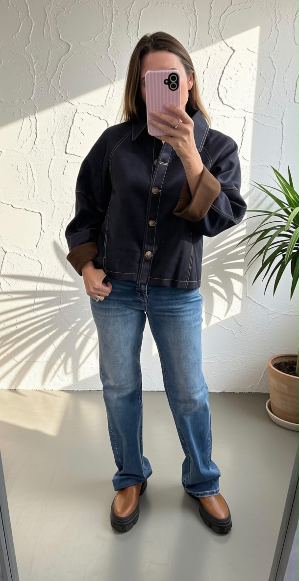 A woman takes a mirror selfie wearing a dark navy blue cropped denim jacket with brown button details and brown cuffs, paired with blue jeans and brown shoes.