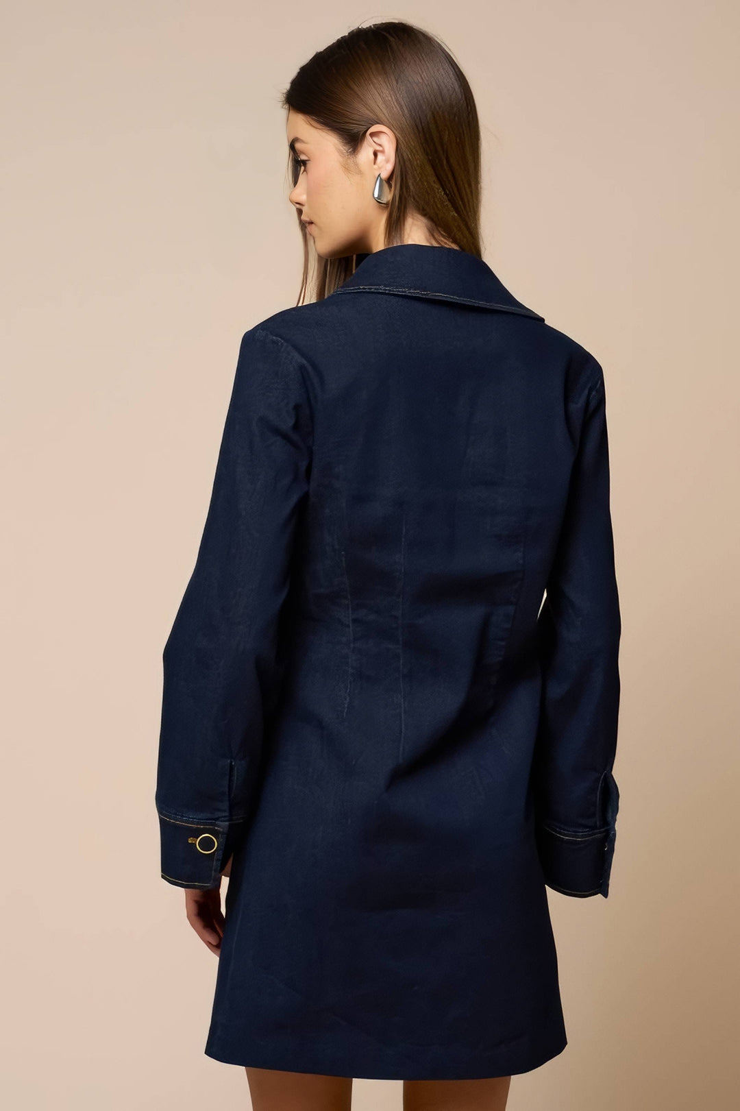 A woman shown from the back wearing a dark navy blue denim-style blazer or coat with a collar and long sleeves, against a plain beige studio background.