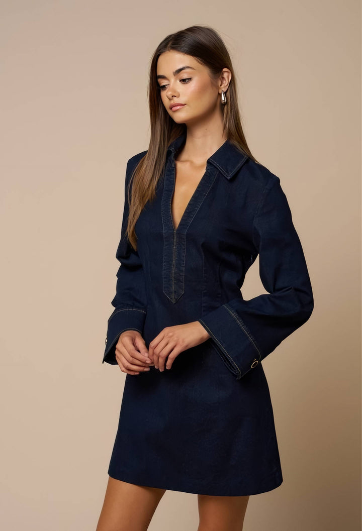 A woman modeling a dark navy blue denim shirt dress with a deep V-neck, buttoned cuffs, and a straight skirt, set against a solid beige studio background.