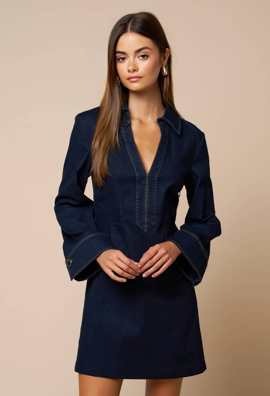 A woman modeling a dark navy blue denim shirt dress with a deep V-neck, buttoned front placket, and flared bell sleeves against a solid beige studio background.