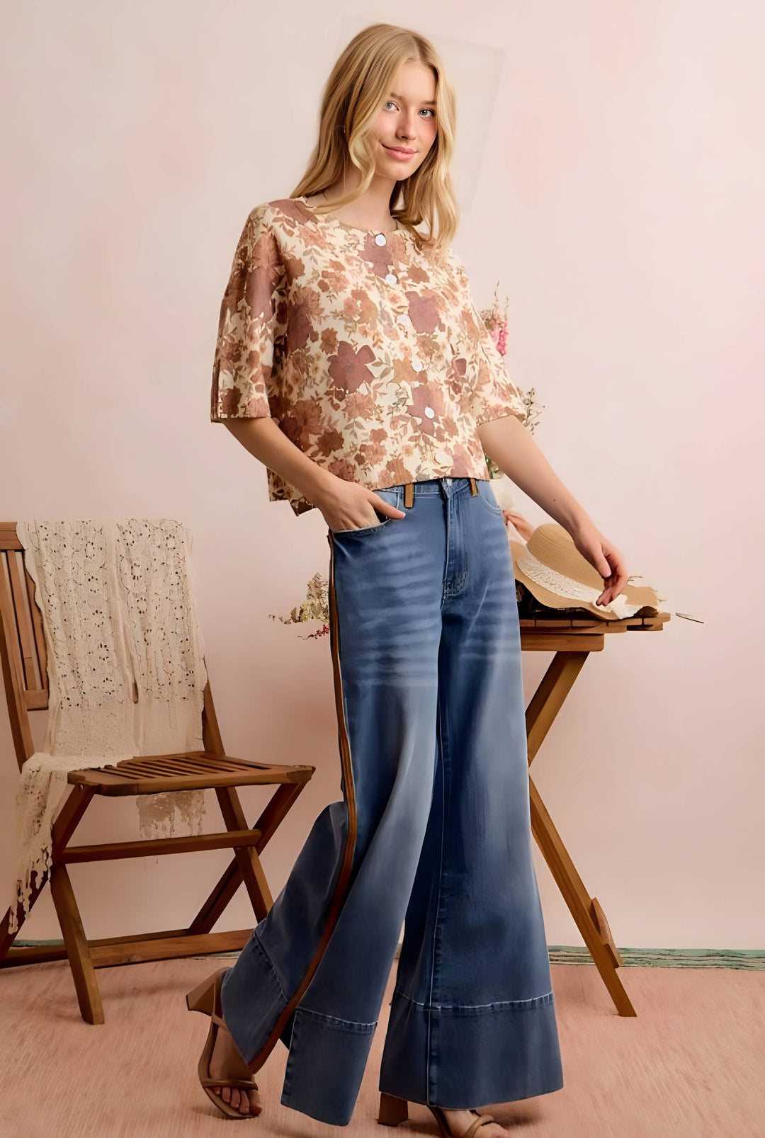 A woman stands in a softly styled room wearing a beige and brown floral short-sleeve blouse with blue wide-leg jeans. She is posed beside a wooden chair and table against a pale pink wall.