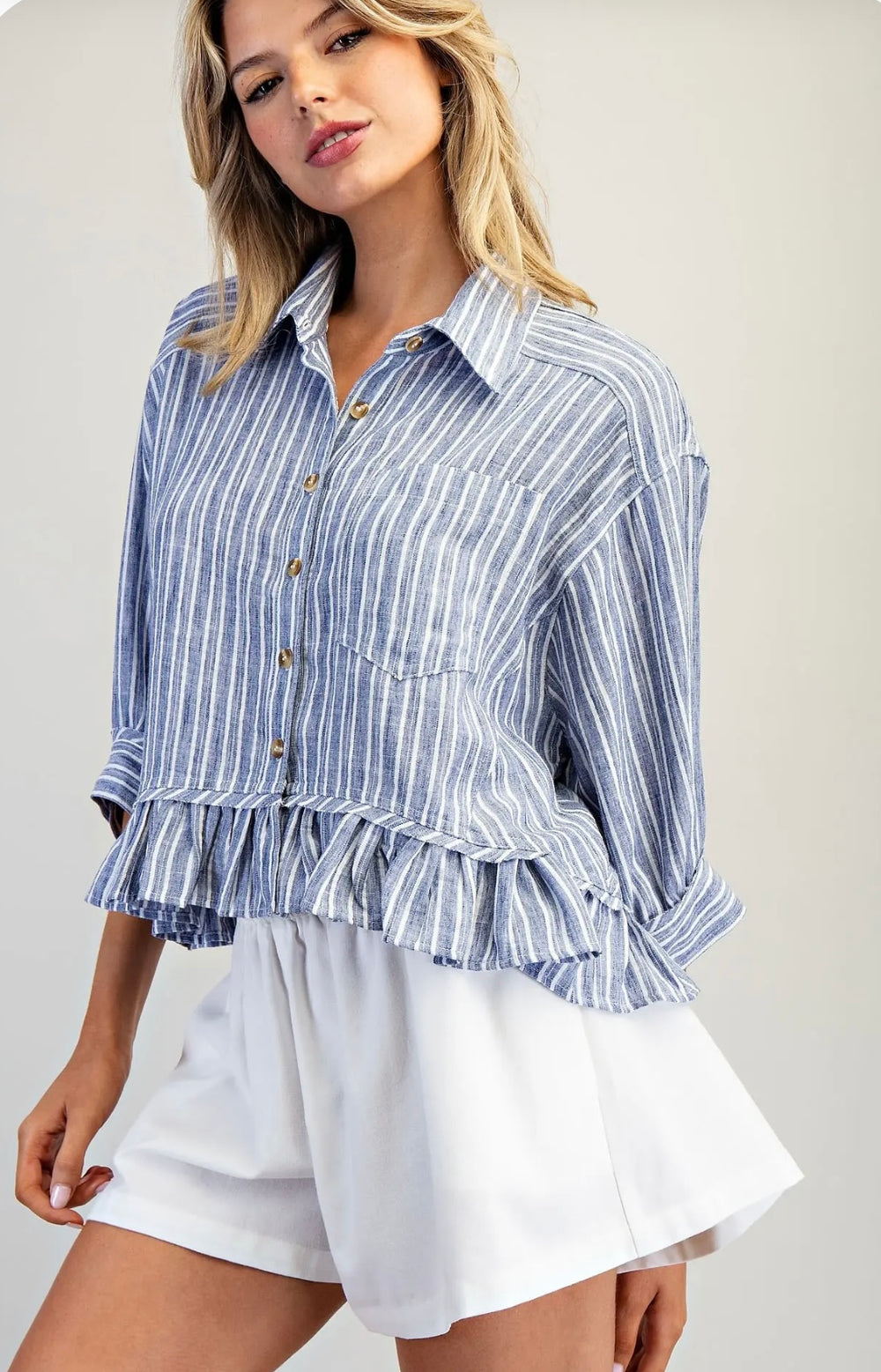 Woman wearing a blue and white striped shirt with ruffled details and white shorts on a plain background