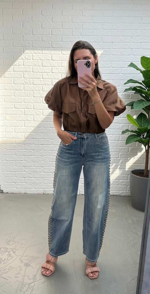 A woman takes a mirror selfie wearing a brown puff-sleeve button-up shirt and high-waisted, wide-leg blue jeans with silver chain detailing down the sides. She is standing in front of a white brick wall next to a potted green plant.