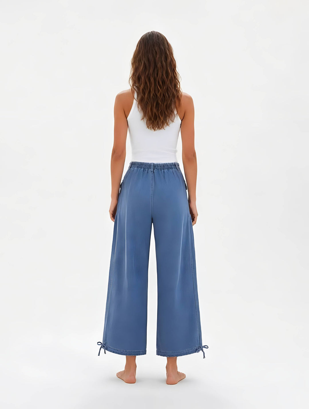 A woman shown from the back wearing wide-leg blue pants with a white tank top, standing against a plain white studio background.