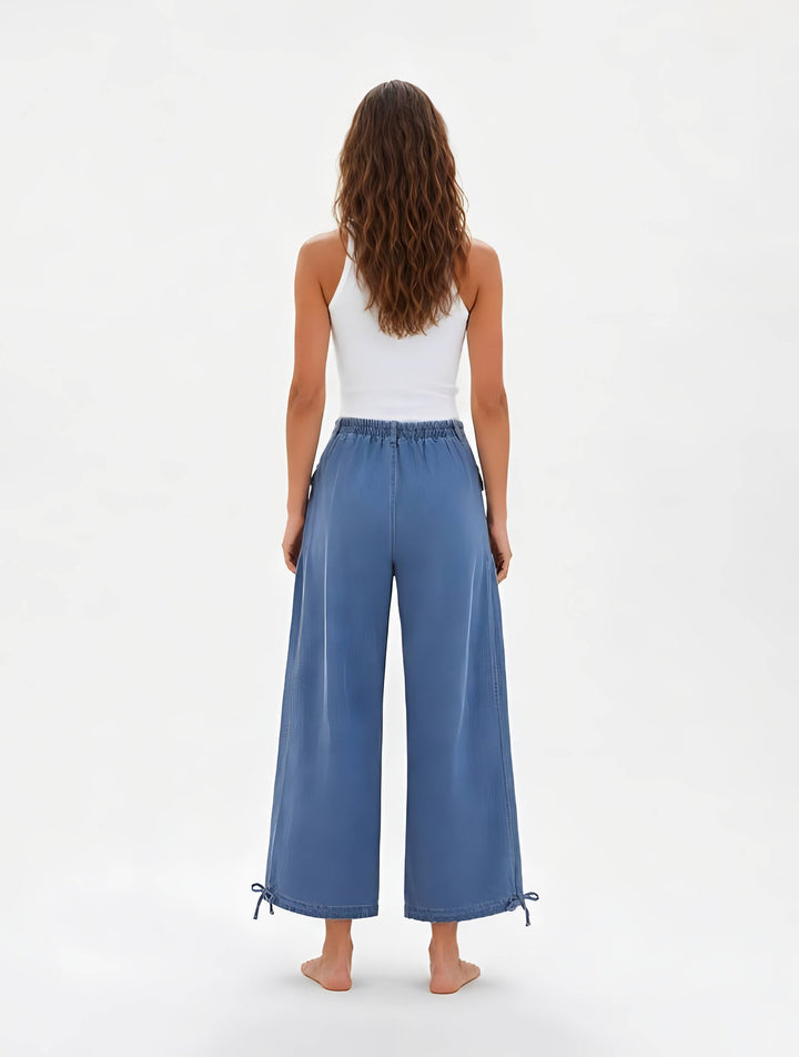 A woman shown from the back wearing wide-leg blue pants with a white tank top, standing against a plain white studio background.