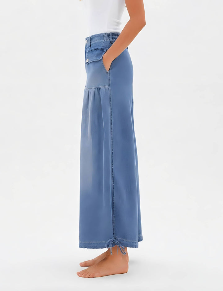 A model wearing a high-waisted, wide-leg blue denim maxi skirt with side pockets and a drawstring hem, shown from the waist down against a plain white background.