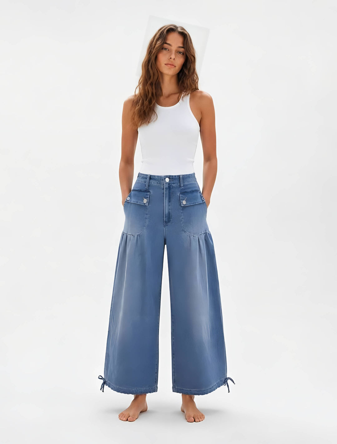 A woman modeling a pair of wide-leg blue denim pants with a white ribbed tank top, standing against a plain white studio background.