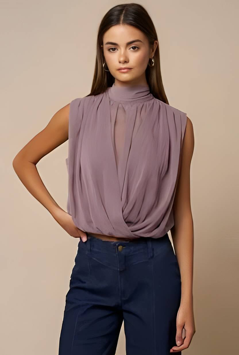 A woman modeling a sleeveless, high-neck, draped mauve blouse paired with dark blue trousers, set against a solid beige studio background.