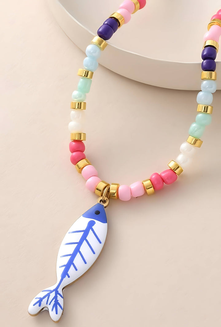 A colorful beaded necklace with a white fish-shaped pendant featuring blue lines. The necklace features pastel pink, light blue, purple, and white beads separated by small gold spacer rings, displayed on a neutral beige background.