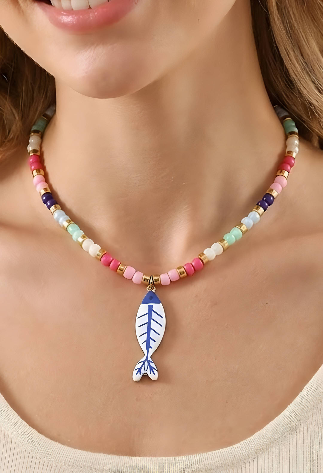 A close-up of a model wearing a beaded necklace with a blue and white fish-shaped pendant. The necklace features pastel-colored beads in pink, blue, green, yellow, and white, separated by small gold spacer beads.