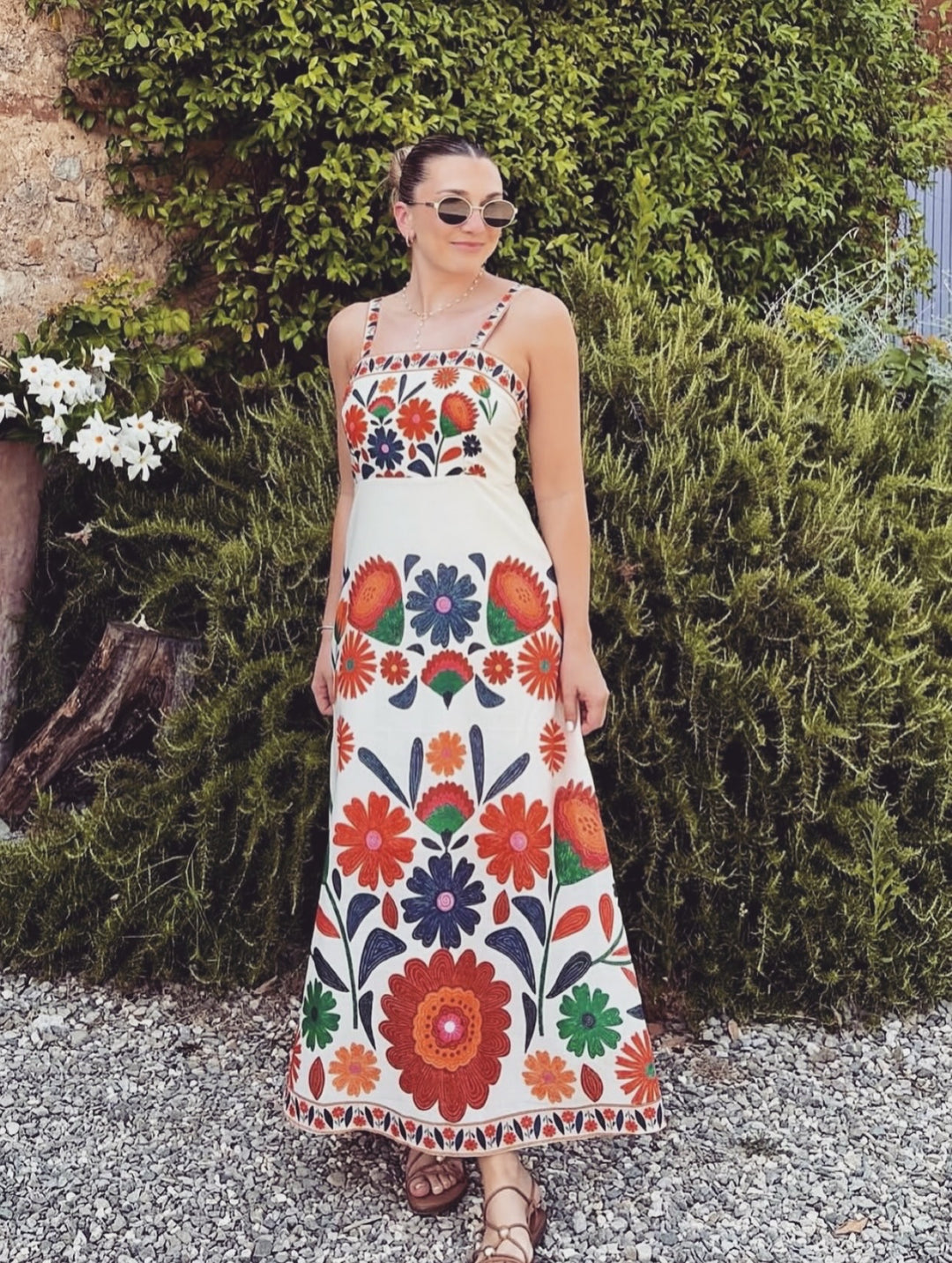 A woman wearing a sleeveless white maxi dress with a colorful floral print, standing outdoors in front of green bushes and a stone wall.