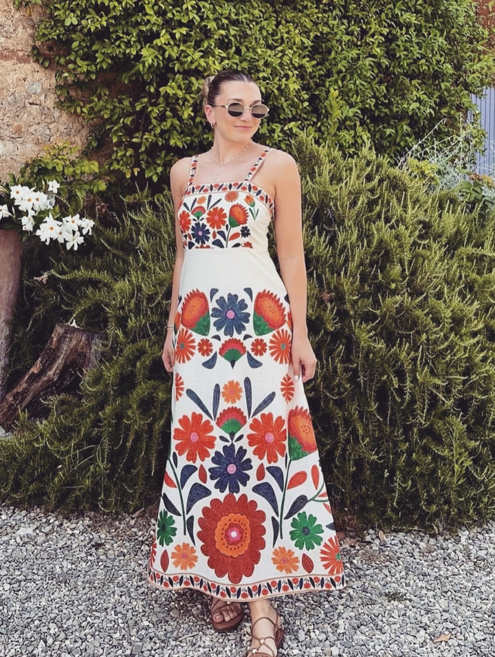 A woman wearing a sleeveless white maxi dress with a colorful floral print, standing outdoors in front of green bushes and a stone wall.