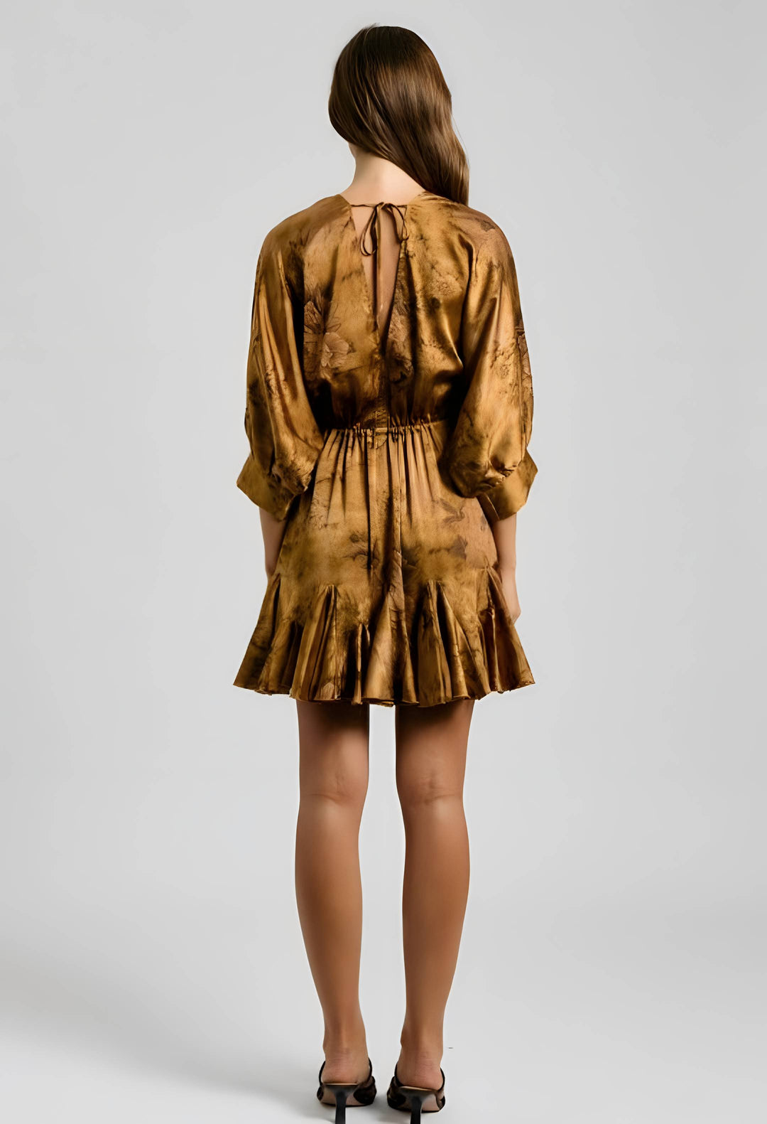 A woman viewed from the back wearing a short metallic gold dress with a tie-dye pattern, three-quarter sleeves, and a pleated skirt, paired with black heels against a light gray studio background.