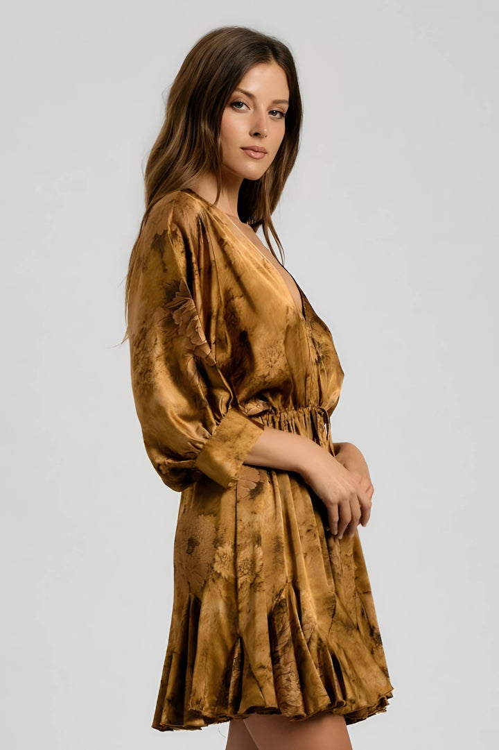 A woman modeling a short, shiny mustard-gold dress with a mottled, marbled pattern in darker brown tones, set against a plain light gray studio background.