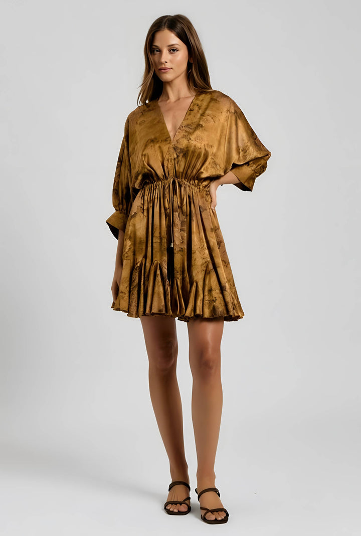 A woman modeling a short, loose-fitting satin-like mini dress in a metallic bronze-gold color with a subtle tonal pattern, paired with black strappy sandals against a light gray studio background.